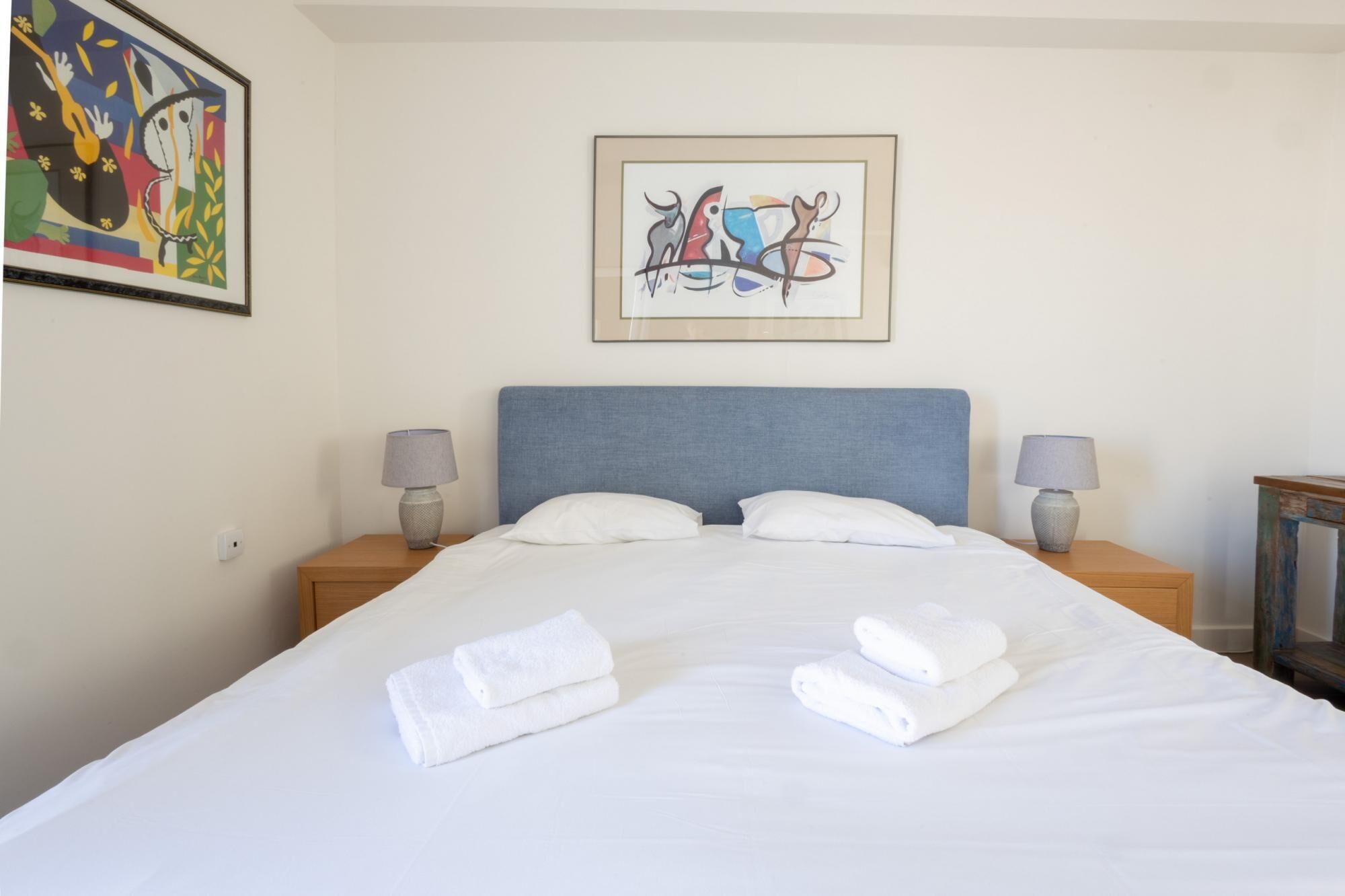 A hotel room with a large bed, two bedside lamps, a framed abstract painting, and a small table with a lamp.