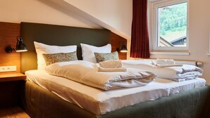 Una cama bien hecha con sábanas blancas y varios almohadones, cabecera de madera, mesita de noche con lámpara y ventana con vista a follaje.