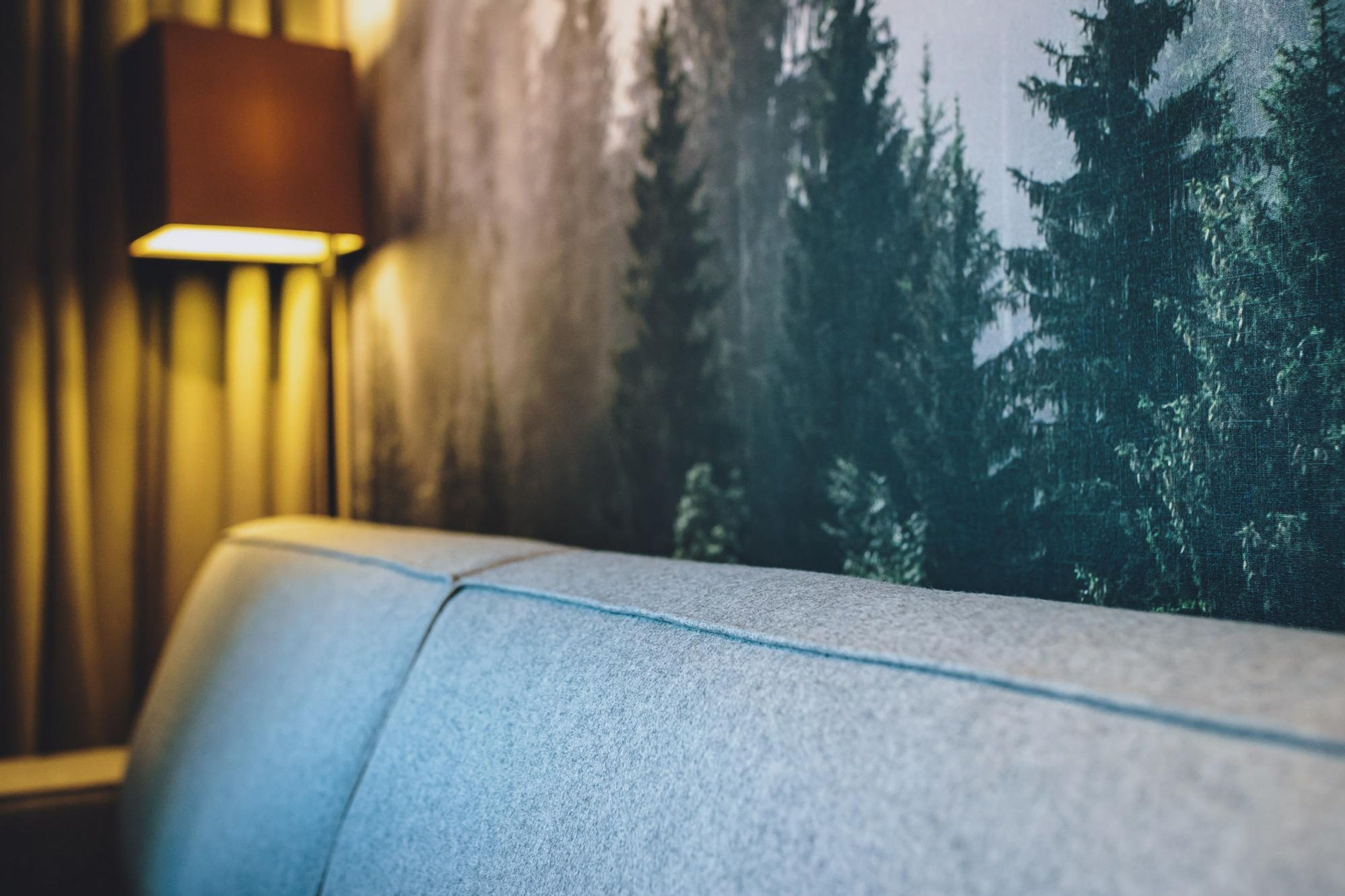 Ein Detailaufnahme eines blauen, gepolsterten Sofas mit sichtbarer Naht, einer Wandlampe und einem Wandgemälde mit Waldmotiv im Hintergrund.