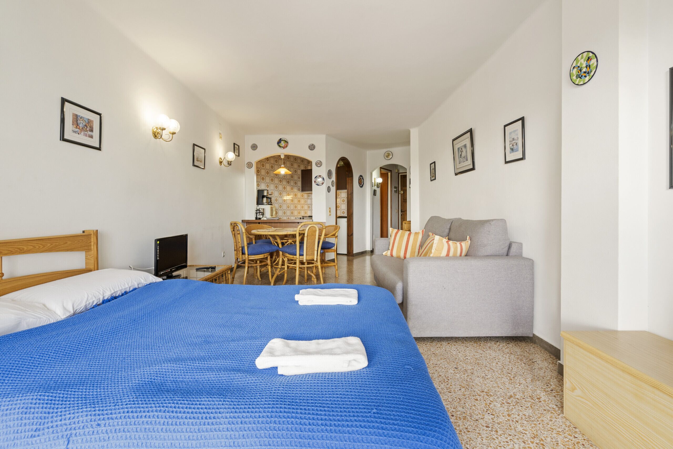 A hotel room with a large bed, a grey sofa, a dining area with a table and chairs, and a kitchenette.