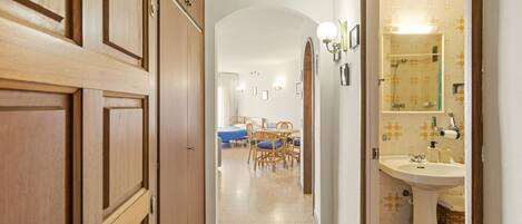 A hallway leading to a bedroom with a bed, a bathroom with a sink and mirror, and a dining area with chairs and a table.