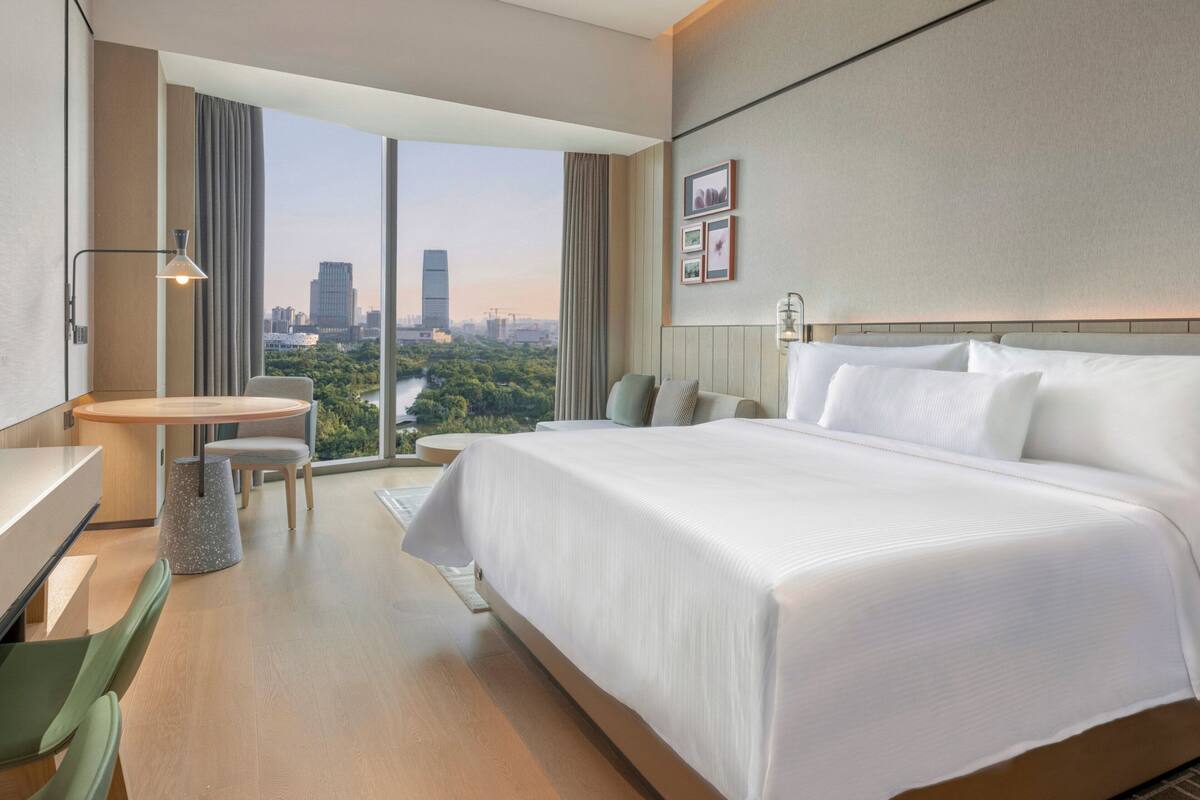 Ett modernt hotellrum med en stor säng, ett skrivbord och en stol. Genom de stora fönstren har man utsikt över stadsbilden.