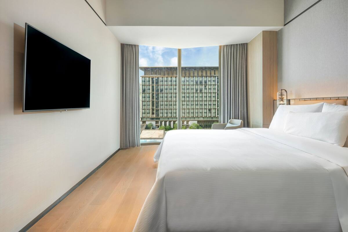 Ett modernt hotellrum med en stor säng, en platt-TV som är monterad på väggen, utsikt över stadsbilden genom stora fönster och en liten sittgrupp med en stol och ett bord.