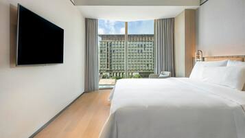 Una habitación de hotel moderna con una cama grande, un televisor de pantalla plana en la pared, vistas de la ciudad a través de amplias ventanas y una pequeña zona de estar con una silla y una mesa.