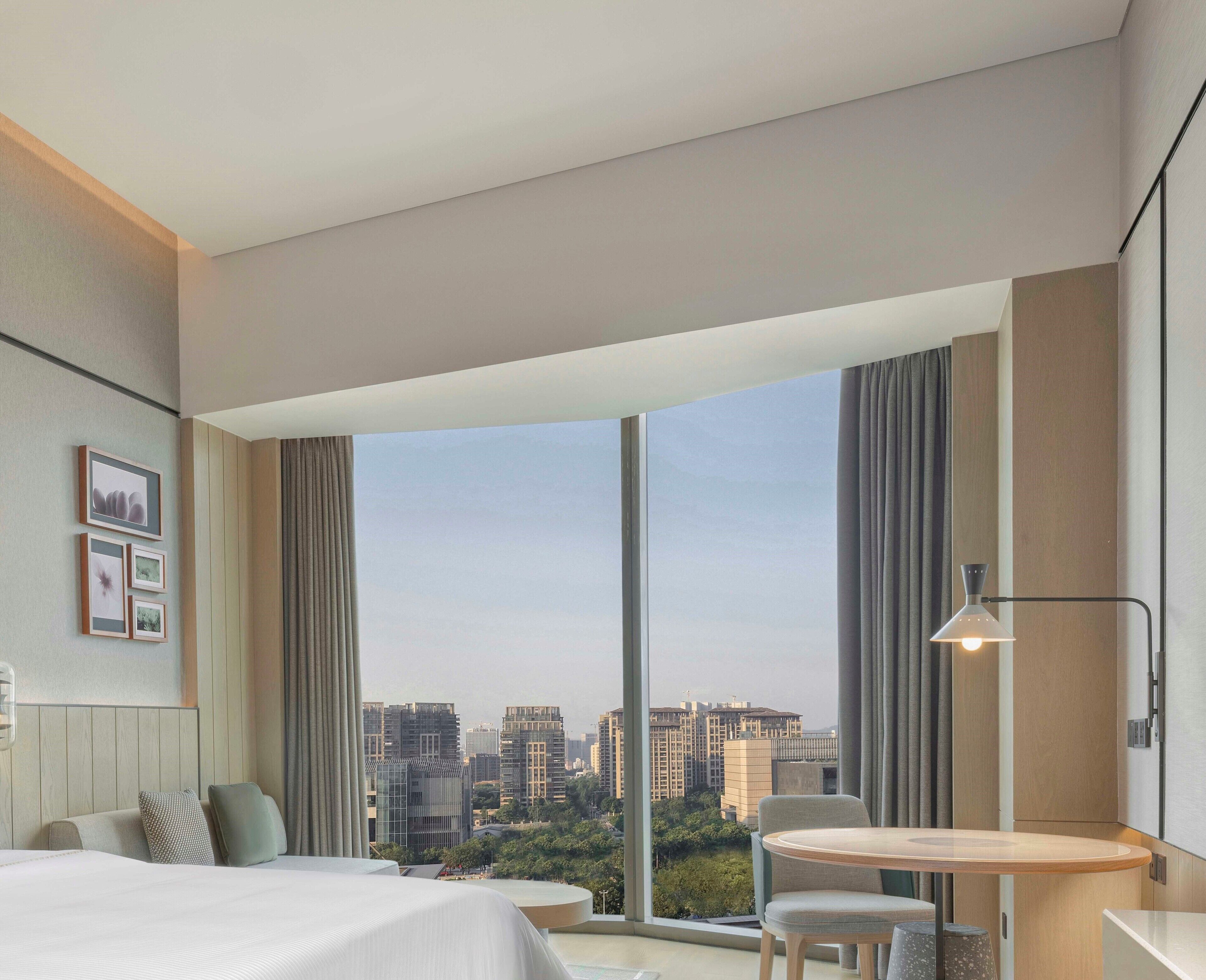 Habitación de hotel moderna con un ventanal grande, una cama, un área de descanso y vistas a la ciudad.