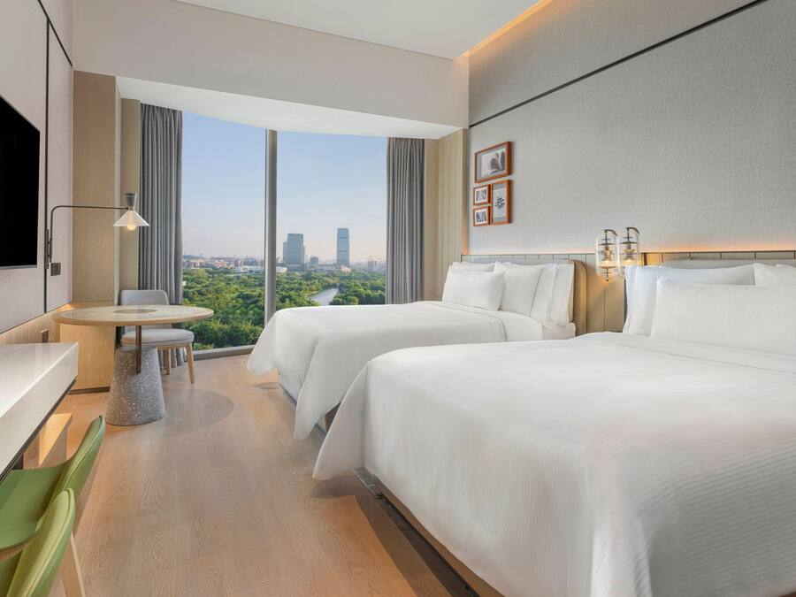 A hotel room with two beds, a desk, a chair, a small table, and a large window offering a city view.