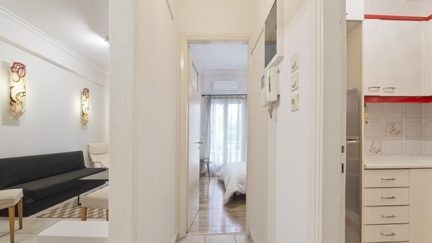 En smal hall leder in till ett sovrum med en säng, ett litet bord och en stol. På höger sida finns ett litet kök med ett kylskåp och skåp.