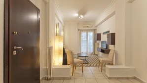 Ett hotellrum med en mörk trädörr, ett element, ett litet bord med stolar, en tv och ett fönster med gardiner.
