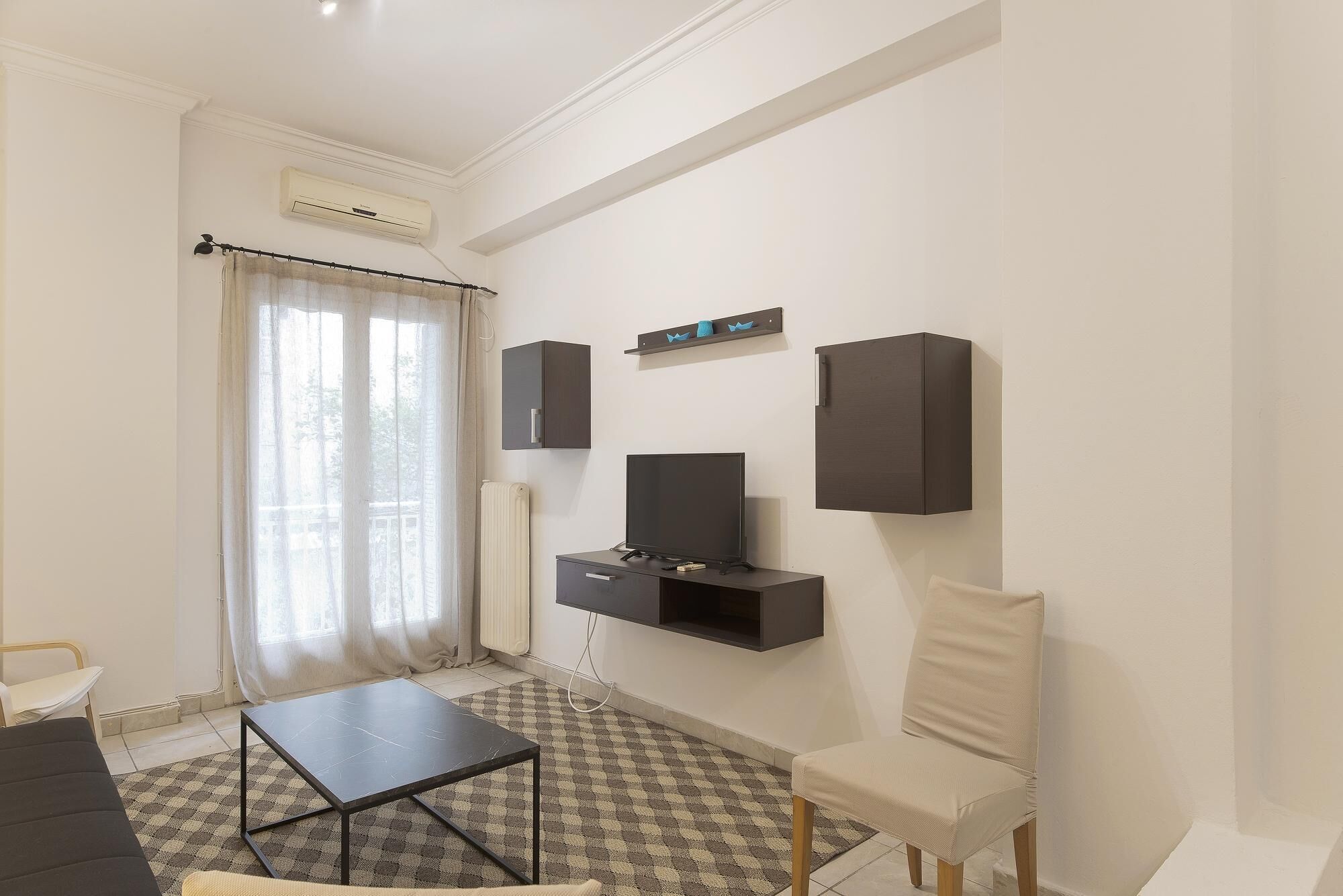 A modern living room with a flat-screen TV, a small black TV stand, a beige armchair, a black coffee table, and a patterned rug.