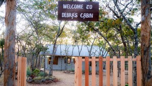 A wooden gate with a sign reading "WELCOME TO DEBRA'S CABIN" leading to a cabin surrounded by trees. - Rustic Luxury Farm Cabin Located in Hwedza - 2031 (Wedza)