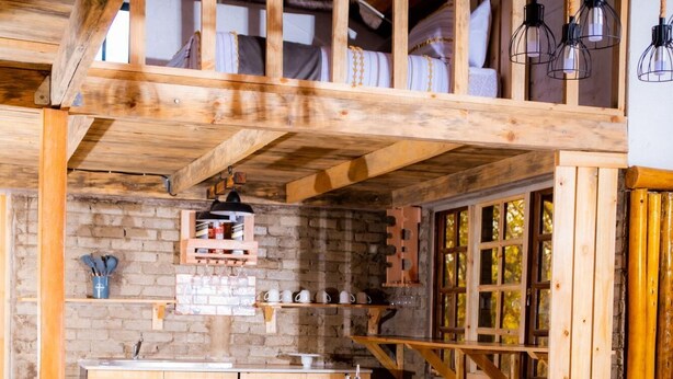 A dining area with a wooden table set for four, surrounded by black chairs. A kitchen with stone walls and wooden cabinets is visible in the background. - Rustic Luxury Farm Cabin Located in Hwedza - 2031 (Wedza)