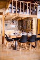 Un comedor con una mesa de madera dispuesta para cuatro personas, rodeada de sillas negras. Se ve una cocina con paredes de piedra y armarios de madera al fondo.