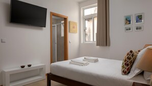 Ein Schlafzimmer mit Bett, Wandfernseher, Fenster mit Vorhängen und einer kleinen Ablage mit Schalen.