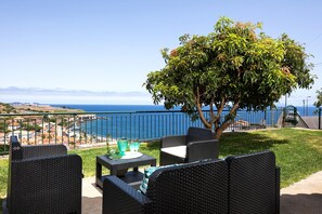 Un área de estar al aire libre con muebles de mimbre, una mesa con mantel individual verde y vistas al mar y edificios lejanos.