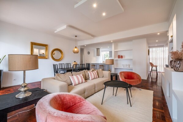 A spacious living room with a sofa, a pink chair, a dining area, and a kitchen in the background.
