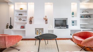 Una sala de estar moderna con un televisor de pantalla plana, dos sillones rosa, una mesa de centro negra y estanterías blancas con objetos decorativos.