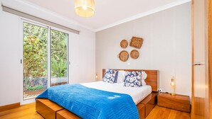 A bedroom with a large bed, wooden flooring, a sliding glass door to a garden, and a wall with woven baskets. - Home to Stay by Homie (Funchal)