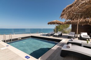 Ein Poolbereich mit Liegestühlen und einem Reetdach-Sonnenschirm mit Blick auf das Meer.