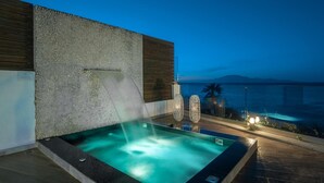 A hot tub with a waterfall feature, surrounded by a stone wall and wooden panel, overlooking a body of water at dusk. - Luxury Villa Cavo Mare Meltemi With Private Pool Jacuzzi (Zakynthos)