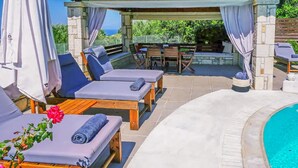 Uma área à beira da piscina com espreguiçadeiras e uma mesa, um espaço de estar coberto e vista para o mar.