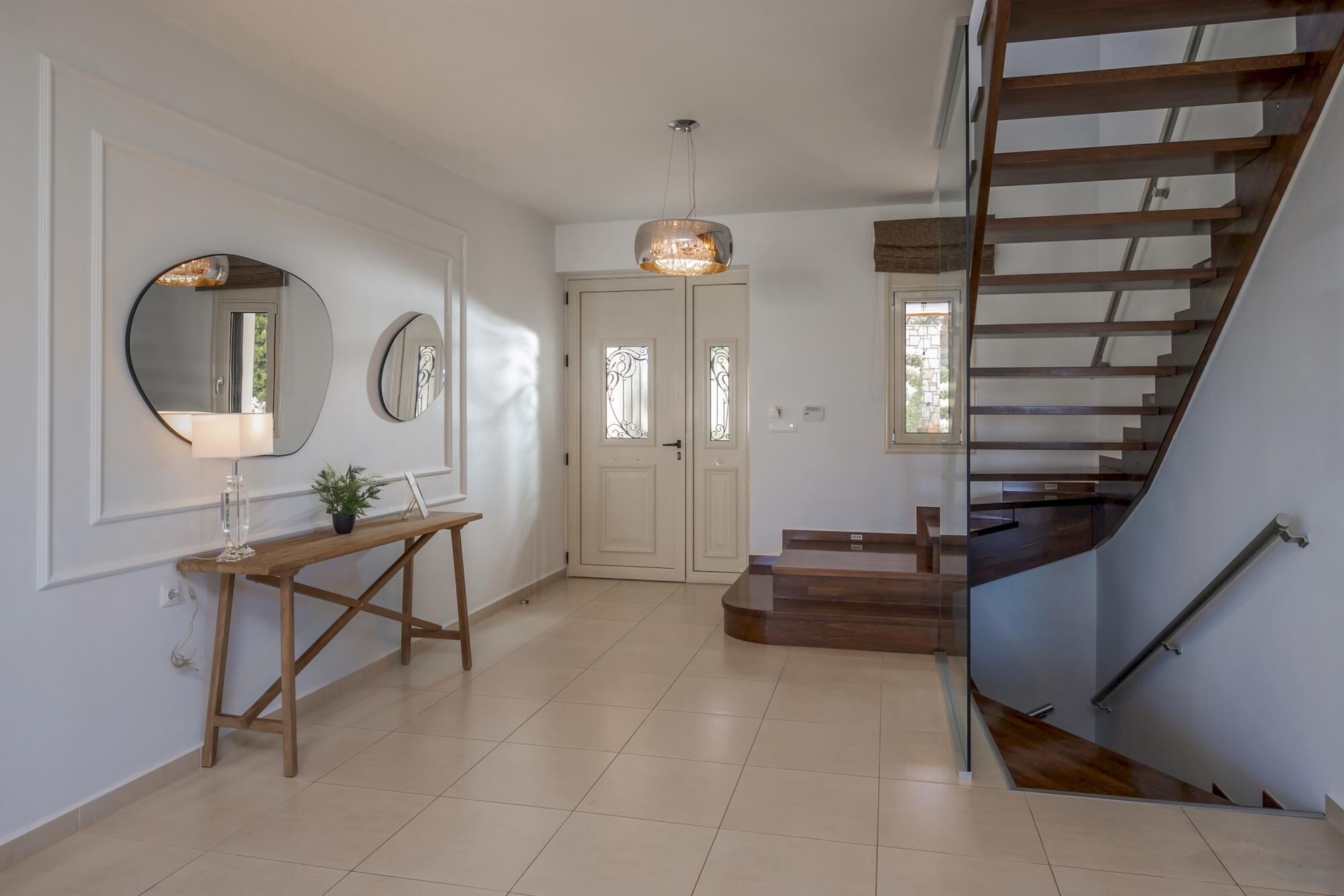 Un intérieur moderne avec un escalier en bois, une console surmontée d’une lampe et des miroirs décoratifs accrochés au mur.