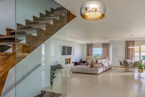 A modern living room with a glass staircase, a large chandelier, a sofa set, and a dining area with a view.