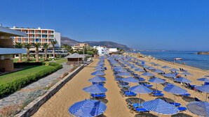 Una spiaggia con file di ombrelloni azzurrati e lettini, un edificio di un hotel e una vista limpida sul mare e sulle montagne sullo sfondo.