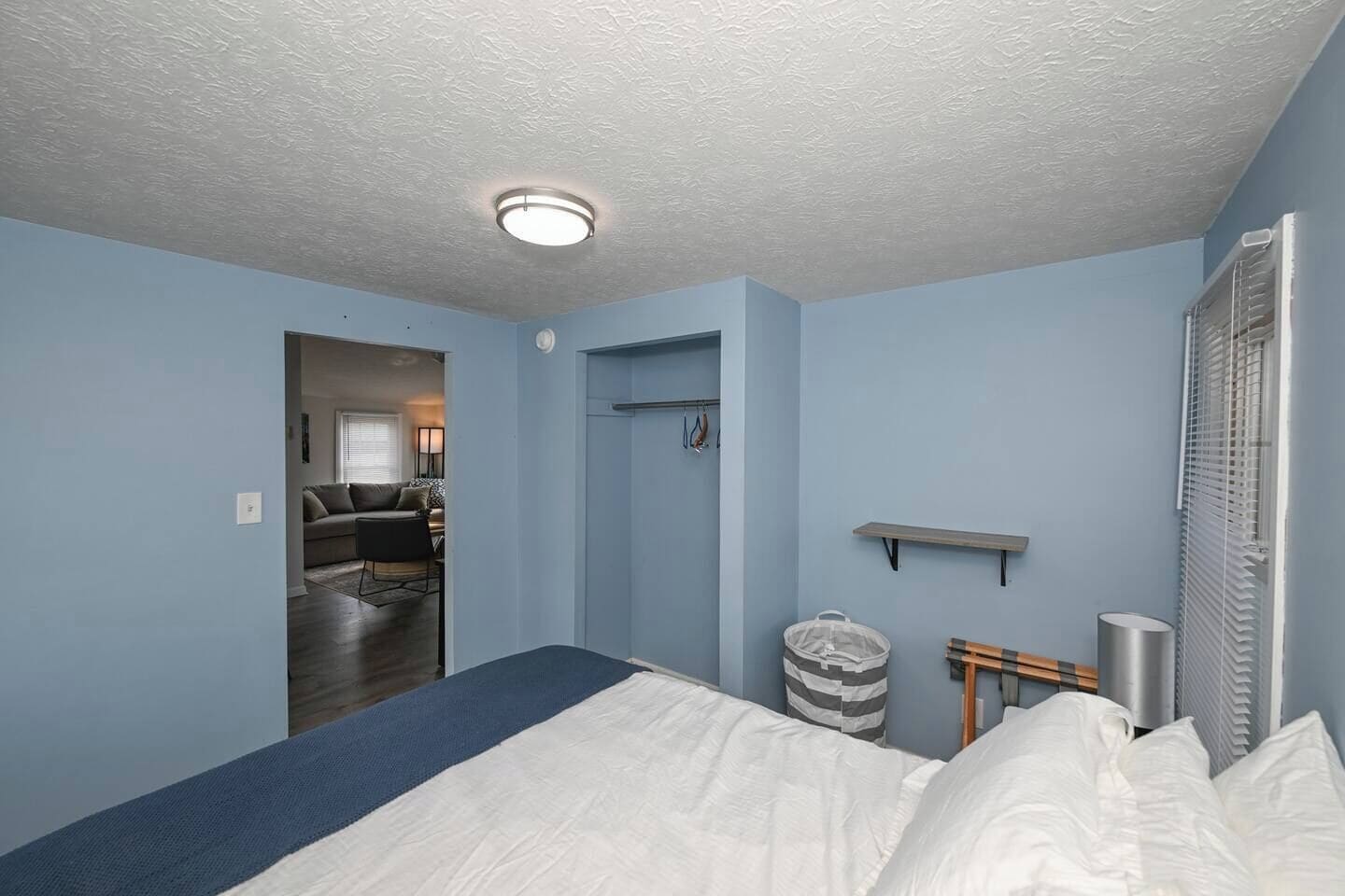 A bedroom with a bed, a small shelf, a trash bin, and a window with blinds.