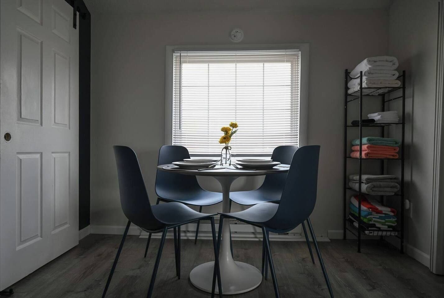 A dining area with a round table, four chairs, a vase with flowers, and a window with blinds.