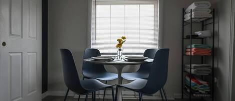 A dining area with a round table, four chairs, a vase with flowers, and a window with blinds.