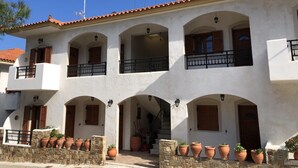 A white building with multiple balconies, stone walls, and terracotta pots. - Nikos Apartments A8 in Gialiskari (Ikaria)