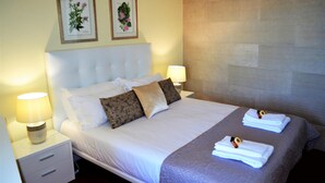Cama bem arrumada com lençóis brancos e duas almofadas, mesa de cabeceira com abajur, e duas gravuras botânicas emolduradas na parede.