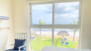 Une chambre avec un lit, un fauteuil à bascule et une grande fenêtre donnant sur une plage parsemée de parasols et de chaises longues.