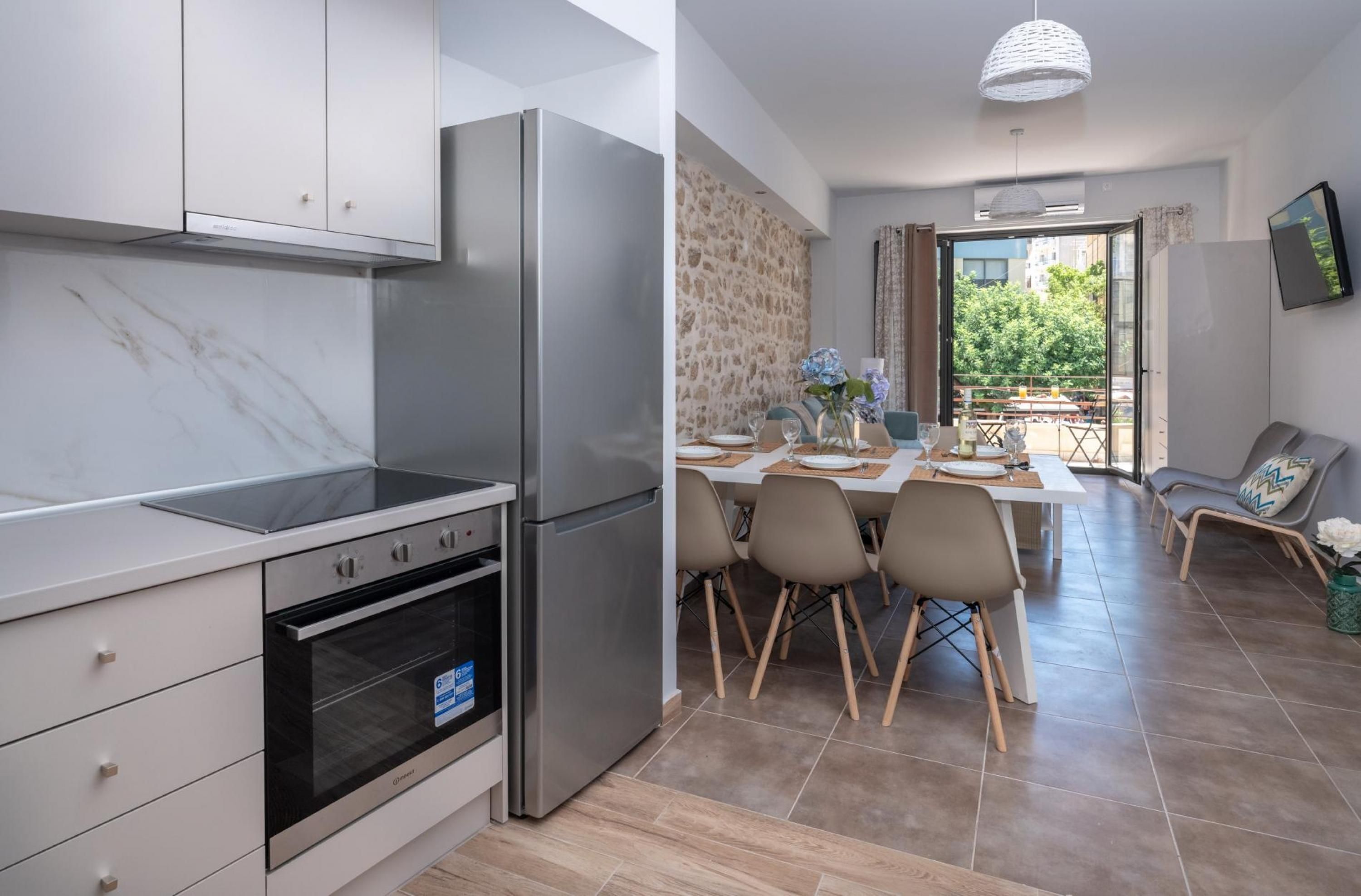 Een moderne keuken met roestvrijstalen apparatuur, een eethoek met een tafel en stoelen, en uitzicht op groen via de open deur.