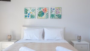 Cama bem arrumada com lençóis brancos e duas toalhas enroladas, com mesas de cabeceira brancas e luminárias de cada lado. Acima da cama, três quadros florais emoldurados na parede.