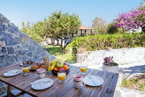 Une table en bois, dressée pour le petit-déjeuner, avec des croissants, des fruits et des verres de jus, le tout sur fond d’un mur de pierre et de verdure.