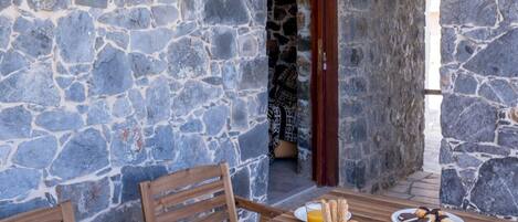 Área de refeições ao ar livre com paredes de pedra e mesa de madeira posta para o café da manhã, com frutas, doces e bebidas.