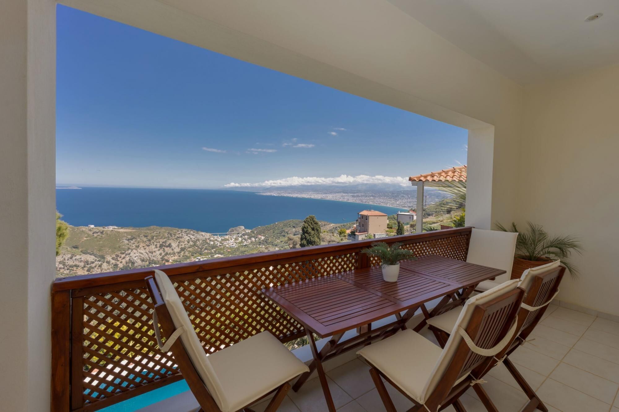 Un balcon avec une table et des chaises en bois, une plante en pot et une vue sur la mer et les bâtiments éloignés.