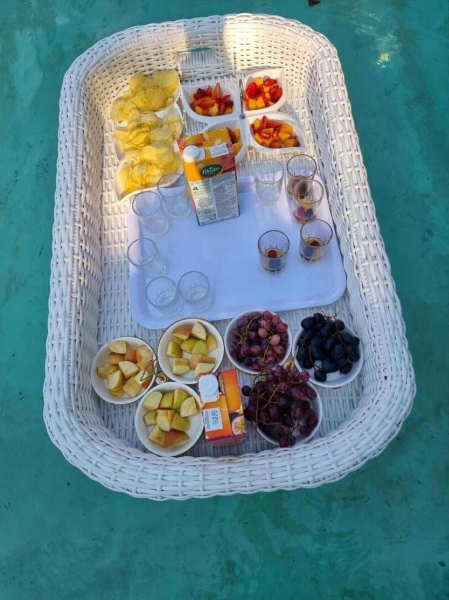 A wicker tray with fruits, a juice box, and glasses on a turquoise surface.