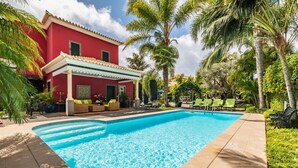 Une piscine dotée d’une terrasse carrelée, entourée de palmiers et bordée d’un bâtiment rouge avec un patio couvert.