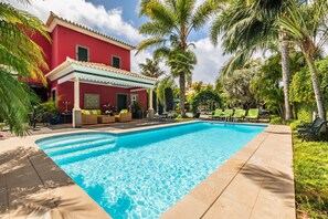Une piscine dotée d’une terrasse carrelée, entourée de palmiers et bordée d’un bâtiment rouge avec un patio couvert.
