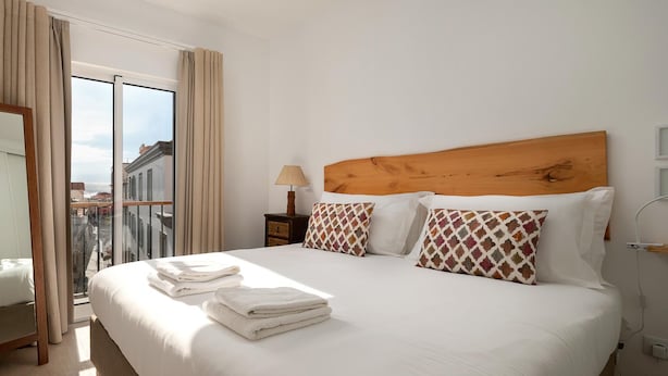 Ein Hotelzimmer mit einem großen Bett, zwei Kissen, einem Nachttisch mit Lampe und Blick auf Gebäude durch das Fenster.