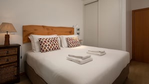 Ein ordentlich bezogenes Bett mit weißen Leinen und zwei gemusterten Kissen, ein hölzernes Kopfteil, eine Nachttischlampe und ein kleiner Nachttisch mit einer Lampe.