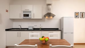 Eine moderne Küche mit weißen Schränken, einer Mikrowelle, einem Toaster und einem Kühlschrank. Ein Holzesstisch mit Stühlen und eine Schüssel mit Obst steht ebenfalls dort.