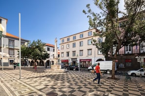 A square with a checkered pavement, a fountain, and buildings with shops.