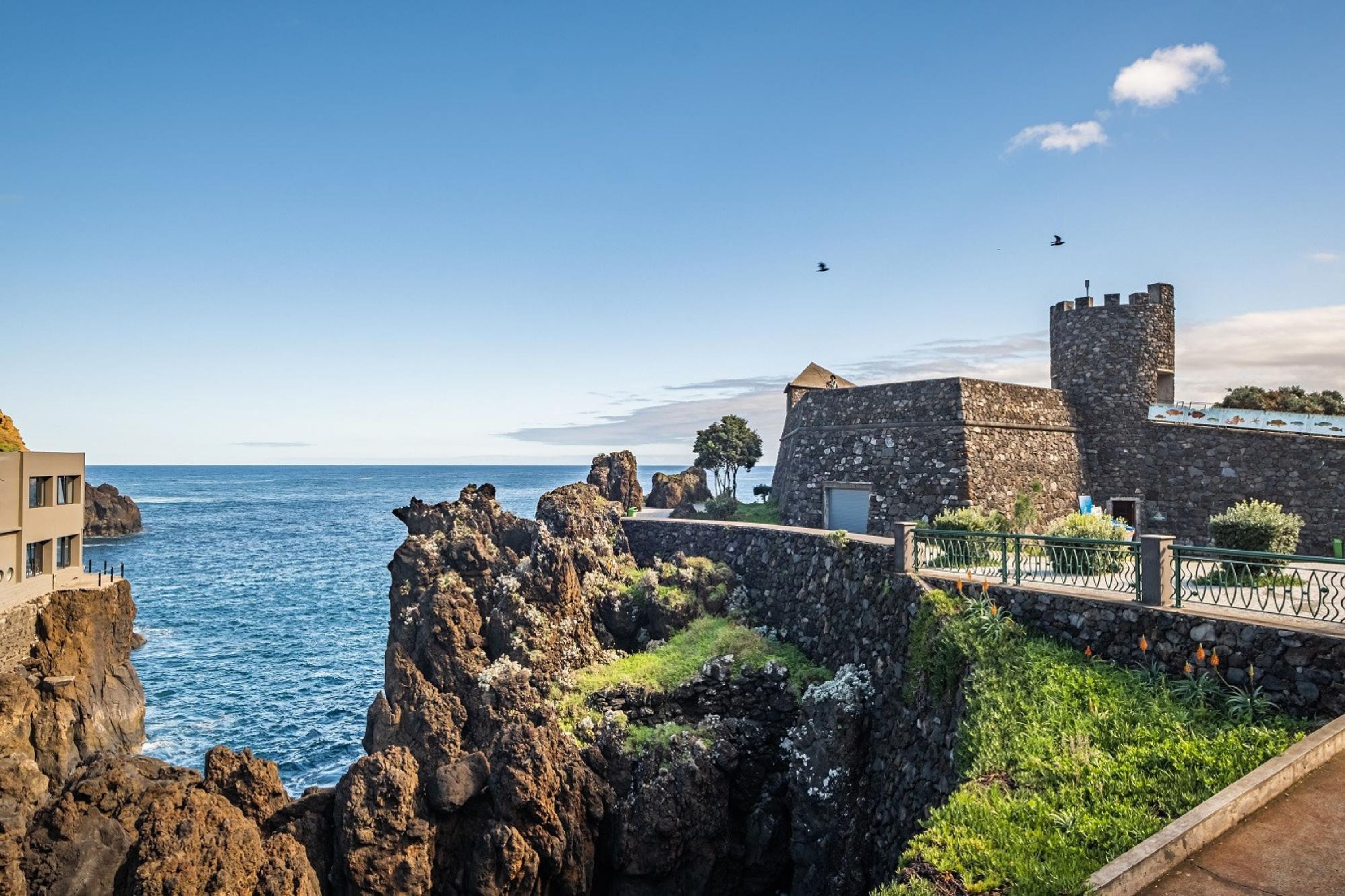 A coastal building with a stone tower, a paved walkway, and a view of the sea.