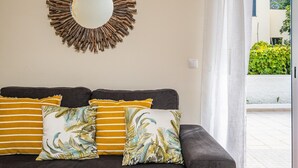 A modern living room with a dark sofa, three patterned cushions, a circular mirror with a natural wood frame, and a view of a green garden through a glass door. - Porto Moniz City Centre by Homie (Porto Moniz)