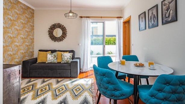 A living room with a sofa, a coffee table, and blue chairs. There is a dining table with a plate of food and drinks. A large window with curtains is visible, and a wall-mounted mirror is present. - Porto Moniz City Centre by Homie (Porto Moniz)