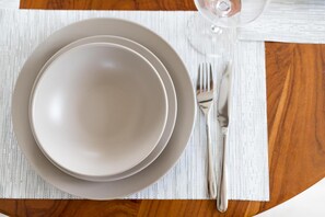 A dining setup with a white placemat, a plate, a bowl, a fork, and a wine glass on a wooden table.