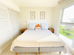 A bedroom with a large bed, two nightstands, a window with a view of greenery, and two wall art pieces.